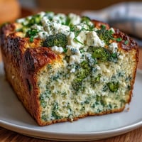 A freshly baked Broccoli and Feta Loaf with golden crust, sliced to reveal moist interior flecks of green and creamy white cheese, served on a rustic wooden board.